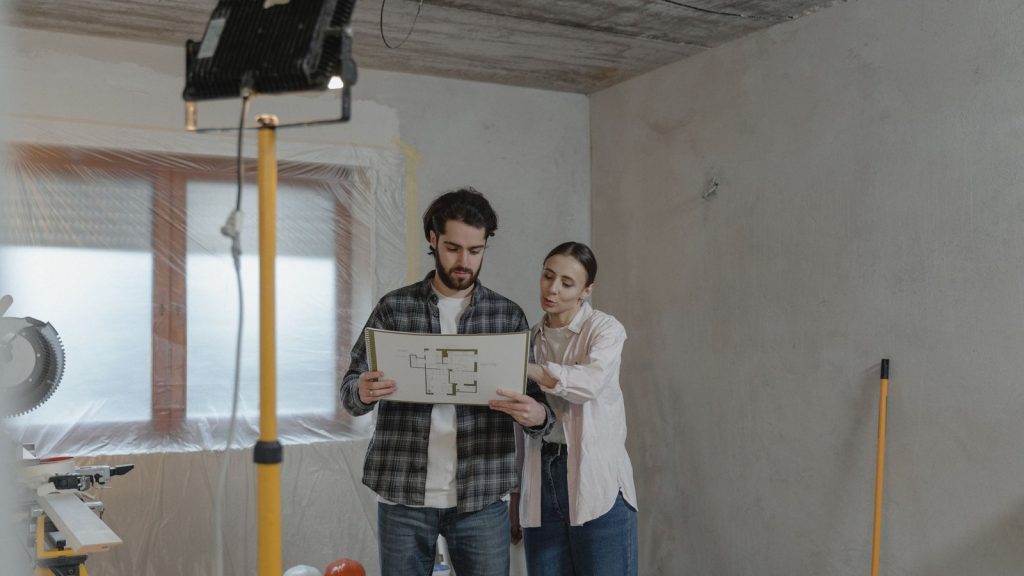 Two adults review a floor plan in an unfinished room during a home renovation.